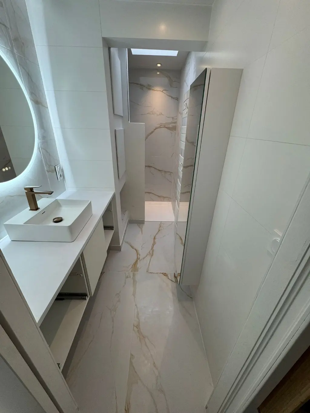 Salle de bain moderne rénovée avec grande douche italienne, carrelage effet marbre et meuble vasque blanc avec robinetterie dorée dans une maison près de Vannes.